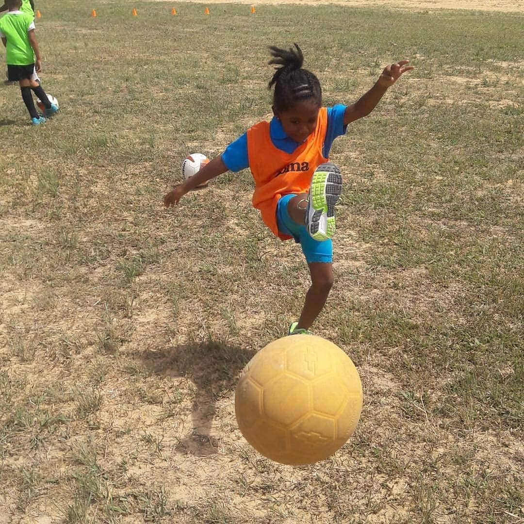 Young girl in an orange bib kicking a football on a dry grass field.