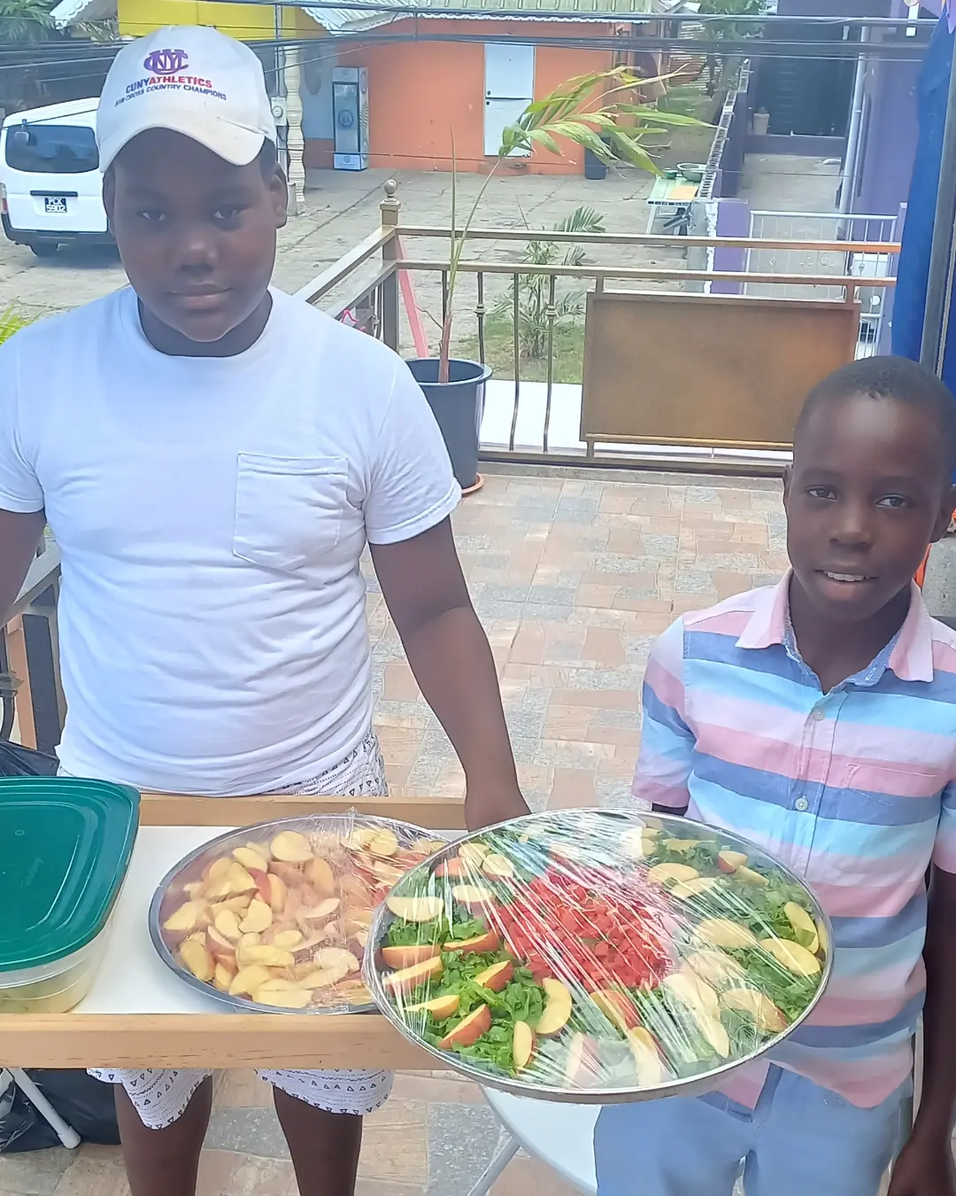 Two boys proudly displaying large trays of prepared fruit salad.