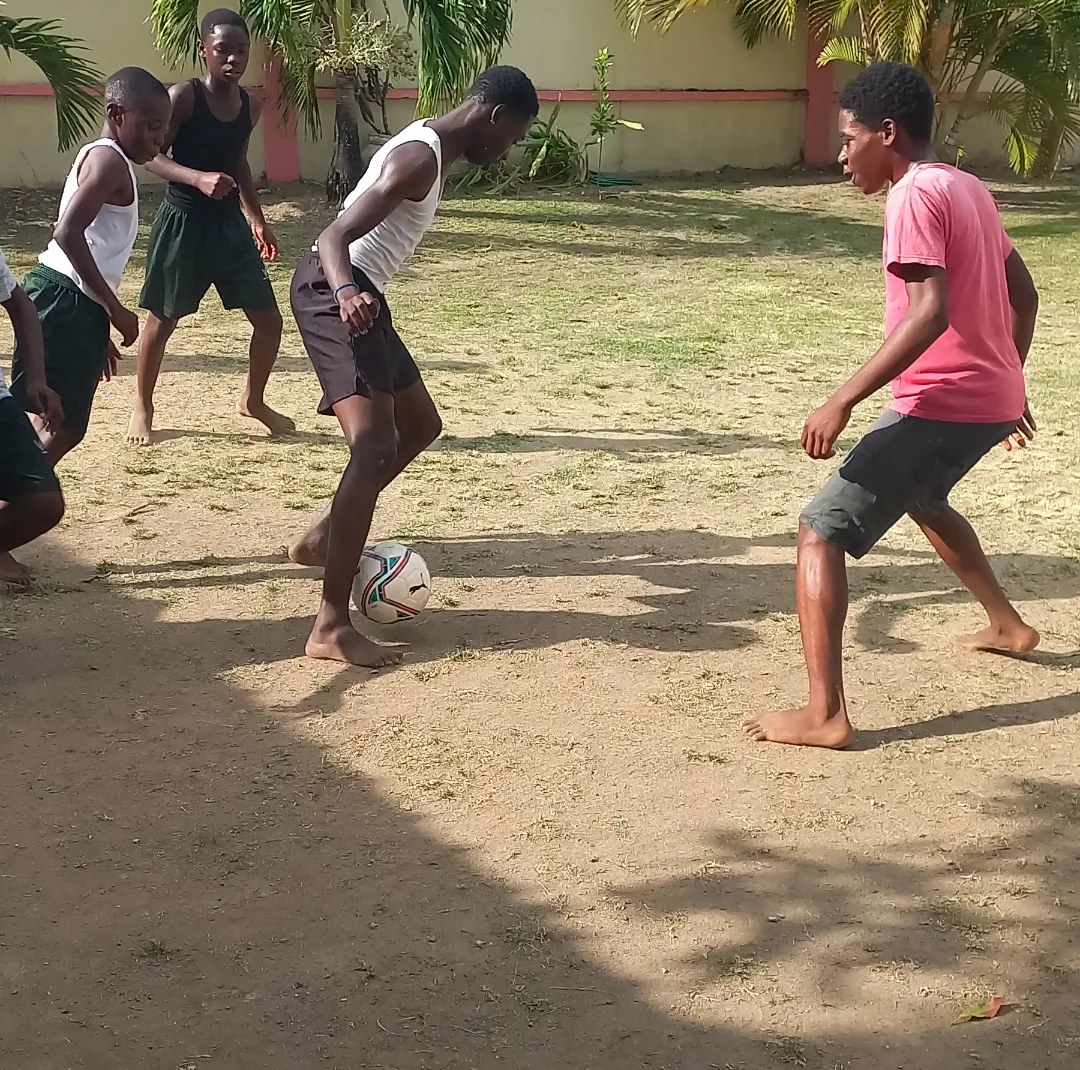Youth playing barefoot football on a sandy pitch, ball at their feet.
